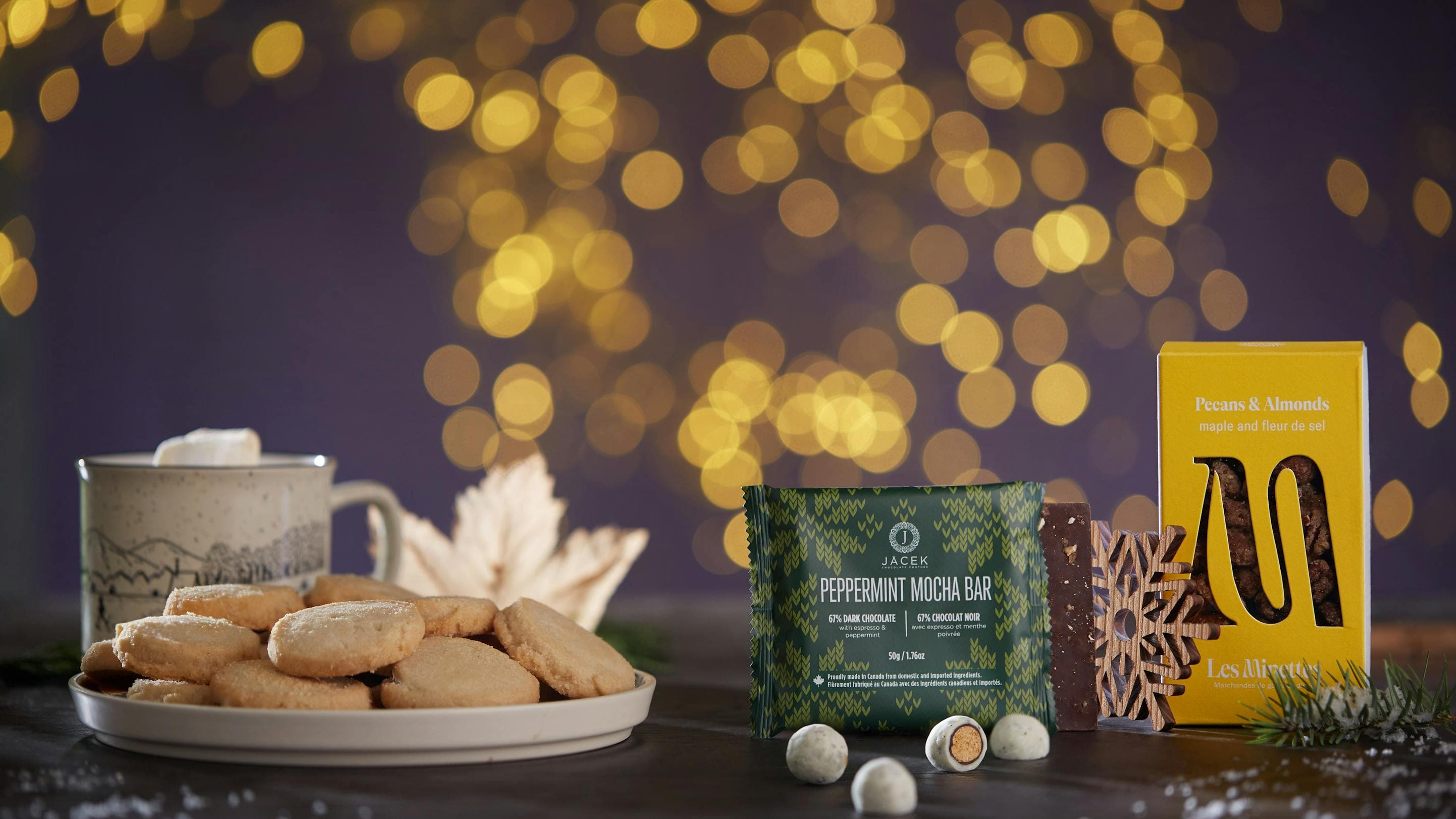 A table full of Christmas treats that include hot chocolate, cookies and chocolate. There are little white lights in the background.