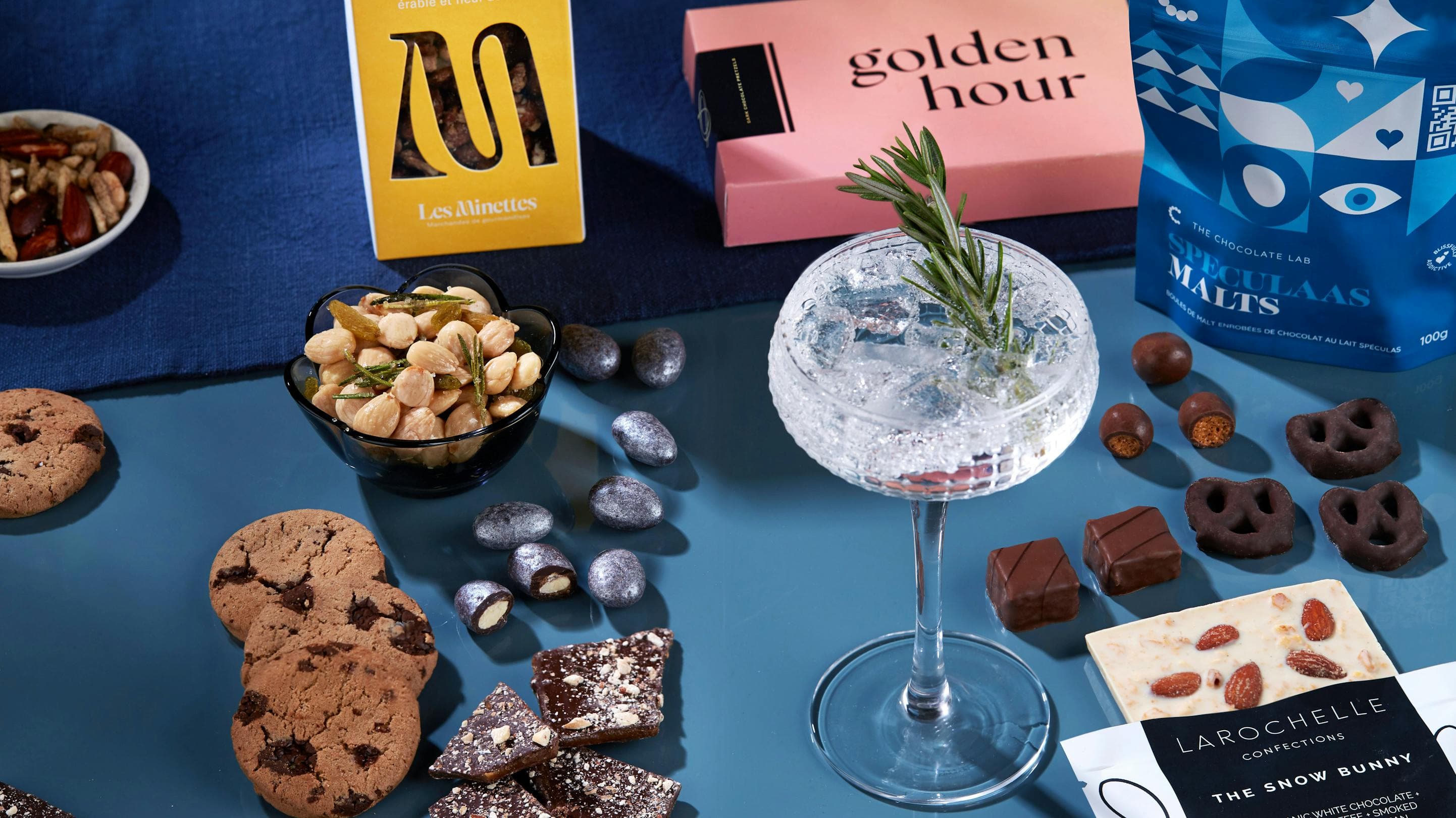 On a blue table, there are cookies, chocolates, mixed nuts, chocolate covered pretzels and a cocktail with a sprig of rosemary.